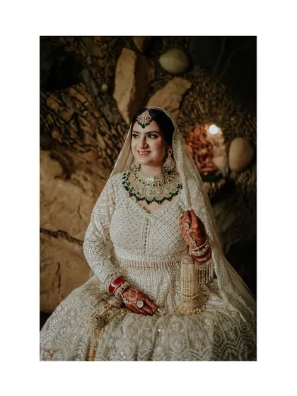 A regal bridal portrait. The bride is seated against a rustic stone wall, her magnificent white and green lehenga spread around her, creating a look of timeless grace and poise.