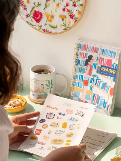 A cozy, reader-themed scene for a stationery brand. The over-the-shoulder perspective invites the viewer into the moment of journaling with a cup of coffee.