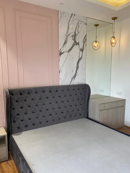The 'before' shot of a bedroom renovation, showing the initial placement of the grey tufted bed against a pink paneled wall and a statuario marble accent.