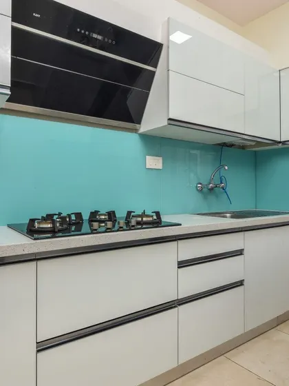 A sleek, modern kitchen with handleless white cabinets and a stunning turquoise glass backsplash. The design includes a built-in hob and chimney for a seamless and minimalist look.