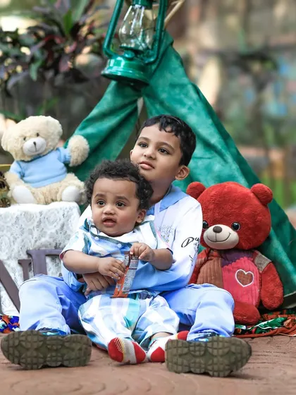 Two brothers sit together for a portrait, with the older one protectively holding his younger sibling. The image, set against their outdoor play tent, highlights their sweet connection.