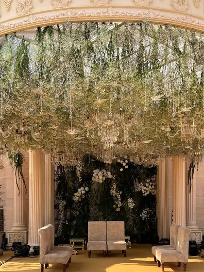 A Roman-inspired mandap featuring grand pillars and a ceiling of cascading green vines and crystal chains. The design combines historical grandeur with the freshness of a secret garden.