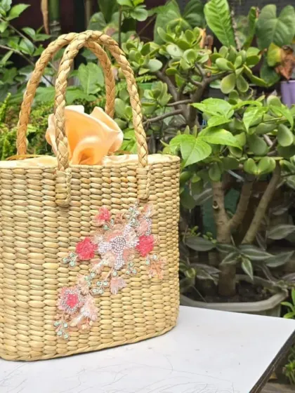 A chic and versatile tote bag, handcrafted from natural grass and adorned with subtle floral embroidery. It features a cloth lining with a drawstring closure, making it both beautiful and practical.