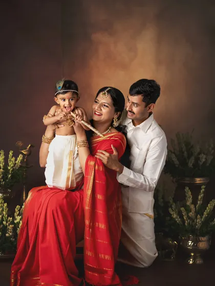 A beautiful family portrait with their little one dressed as Krishna. The traditional outfits and the baby's playful interaction with the flute create a culturally rich and heartwarming image.