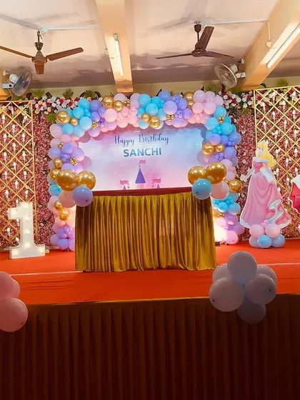 A dreamy Disney Princess themed stage for a first birthday. The decoration features cutouts of various princesses like Ariel and Cinderella, a pastel balloon arch, and a backdrop with a castle silhouette.