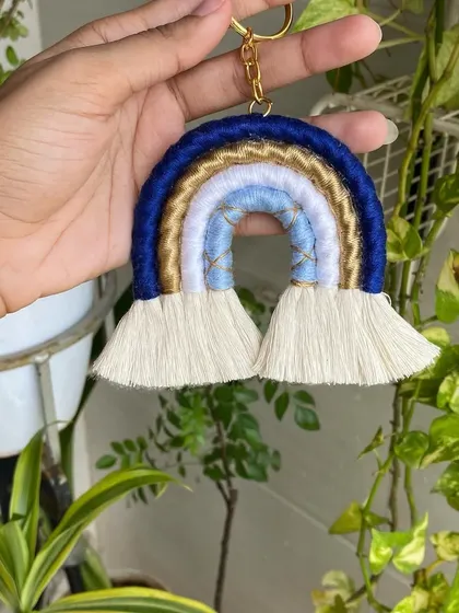 A hand holding our blue and gold macrame rainbow keychain. The natural background highlights the vibrant colors and handmade texture of the piece.