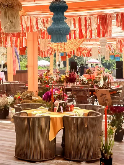 A dining table at the Pahadi Mehendi, with rustic rope-woven chairs and a colorful tassel chandelier hanging overhead, creating a vibrant and textured look.