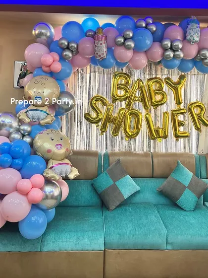 A clear, straight-on view of the at-home baby shower setup, with pink, blue, and silver balloons framing a "BABY SHOWER" sign above a comfortable living room sofa.