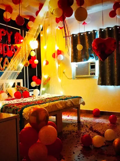 A romantic bedroom setup featuring a canopy with fairy lights, red and white balloons, and a "Happy Birthday" foil banner. The floor is scattered with balloons, creating a fully immersive, love-filled space.