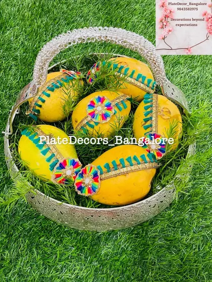 Fresh yellow mangoes decorated with colorful threadwork and ribbons, presented in a silver basket. This design is perfect for summer weddings and functions.