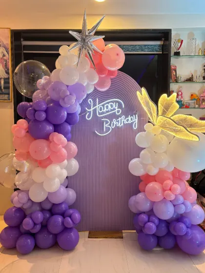 A beautiful pastel butterfly-themed backdrop with a ribbed arch panel, neon sign, and a mix of purple, pink, and white balloons.