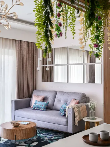 A cozy corner of a living room, featuring a grey sofa, a round wooden center table, and hanging plants that add a touch of nature. The large mirror wall makes the space feel larger.