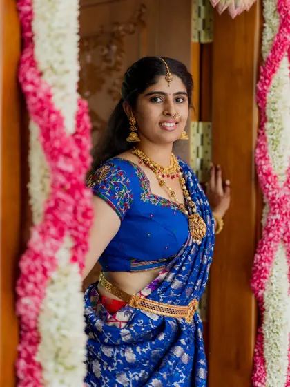 A portrait of a beautifully dressed family member at a celebration. Her vibrant blue saree and traditional jewelry stand out against the floral door frame, capturing the festive spirit of the day.