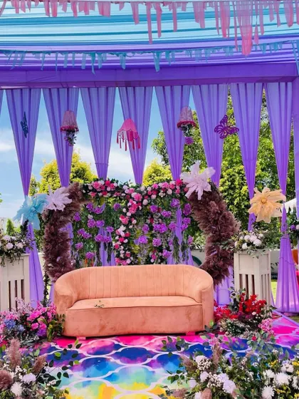 The main stage of a lavender-themed Haldi ceremony. A simple sofa is set against a backdrop of purple drapes and a beautiful floral arch.