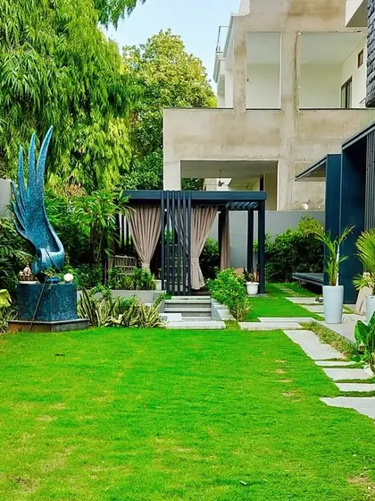 The Jaipur villa's backyard, featuring a lush lawn, a stone path, a striking blue sculpture, and a secluded pergola in the distance.