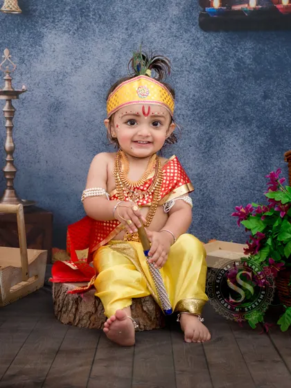 This full-length studio shot captures a smiling baby boy in a complete Krishna costume. The setup includes traditional elements like baskets and diyas against a textured blue backdrop for a rich, festive feel.