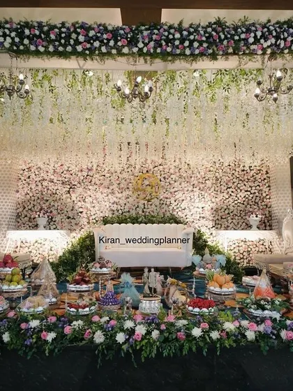 A grand engagement stage decorated with purple and white flowers, hanging chandeliers, and an elaborate display of traditional sweets and gifts.
