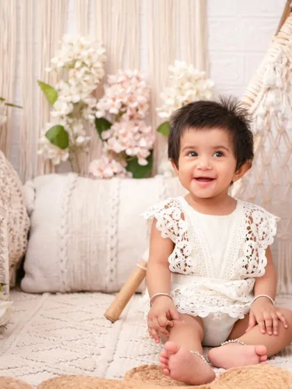 A happy baby girl in a lace romper, perfectly matching the boho-chic aesthetic of the macrame and wicker props.