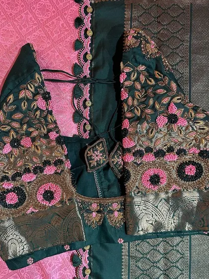 This dark green blouse features beautiful floral embroidery using pink and gold thread, with black beadwork for contrast. The design is complemented by a delicate scalloped border with tassels.