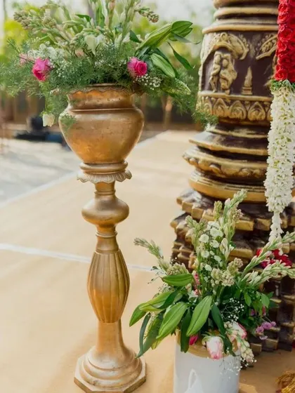 We pay close attention to the details that honor tradition. This image shows a classic brass vase with flowers, set beside a temple-style pillar and a red and white floral garland.