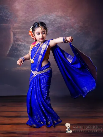 A graceful pose from a young girl wearing a stunning royal blue saree. The simple, elegant backdrop allows her beautiful traditional attire and confident posture to be the focus.