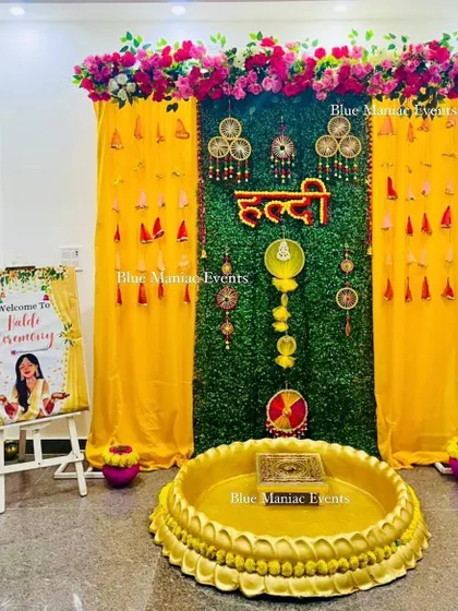 A festive Haldi setup with a green backdrop, yellow drapes, and traditional hangings. The design includes a welcome sign and a central urli for the ceremony.