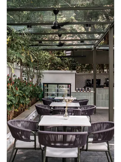 The outdoor dining space at Kana Cafe, surrounded by lush greenery. The seating is covered by a glass canopy, allowing patrons to enjoy the outdoors in comfort, reinforcing the nature-integrated theme.