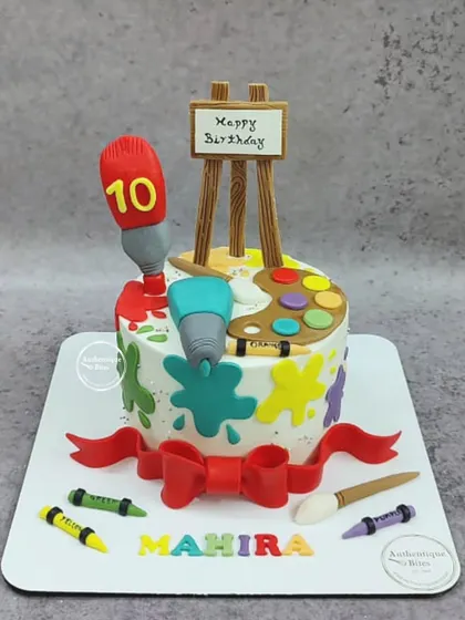 A colorful cake for a little girl who loves to paint. The cake is decorated with paint splatters, an easel, a palette, brushes, and crayons.