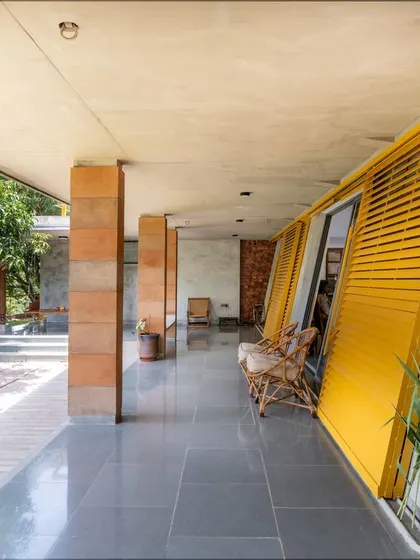 The deep verandah or 'Jagli' at "Aamra" is defined by an inclined wall and bright yellow louvered shutters. This semi-open space provides shade and a seamless transition between the interior and the mango grove outside.
