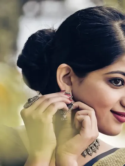 A close-up portrait capturing a moment of getting ready. The focus is on the expression and the traditional jewelry, complementing a classic red saree.