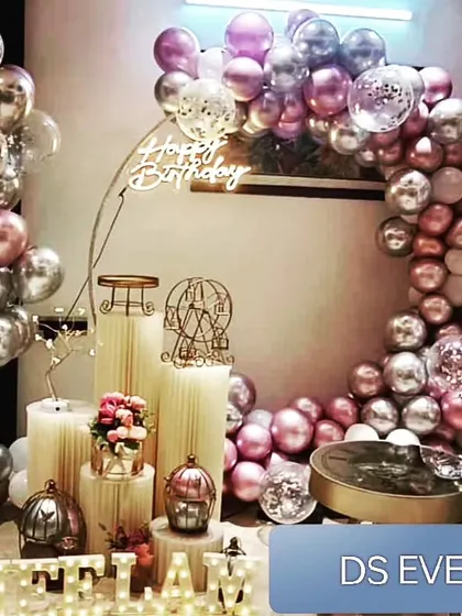 A luxurious party setup featuring a metallic balloon arch in shades of rose gold and silver. The scene is set with elegant white plinths of varying heights, a neon sign, and decorative props like a miniature Ferris wheel.