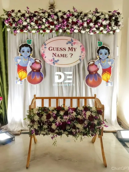 A sweet and simple Krishna-themed cradle decoration for an intimate naming ceremony. The setup includes a floral-draped cradle, a "Guess My Name?" sign, and cute cutouts of Little Krishna.