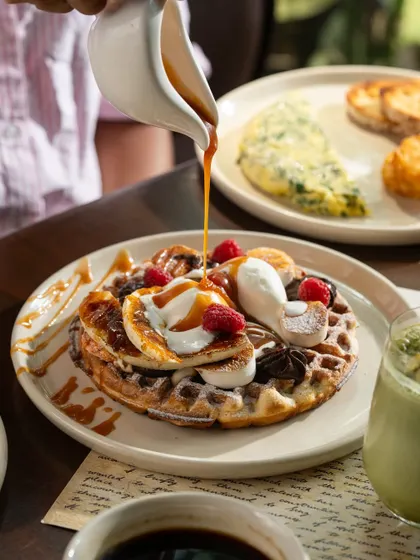 A decadent waffle topped with ice cream, fruit, and caramel sauce. The pouring action adds a sense of indulgence.