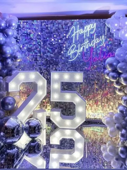 A glamorous 25th birthday backdrop featuring a silver sequin wall that sparkles and shines. The setup is completed with a custom neon sign, marquee numbers, and a silver balloon garland.