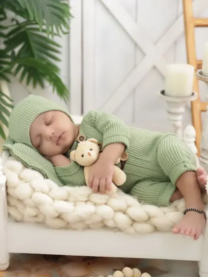 A full view of the cozy setup, showing the newborn in a green knit outfit sleeping on a miniature bed with a teddy bear. The background props add to the rustic charm.