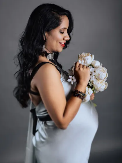 A close-up profile shot of a smiling mother-to-be in a silver saree, holding a bouquet of white flowers. The focus is on her happy expression and the gentle way she holds the bouquet.