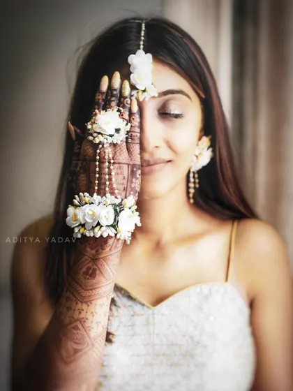 A creative portrait of a bride on her haldi day. She covers one eye with her henna-adorned hand, which is decorated with beautiful floral jewelry, creating a unique and artistic shot.