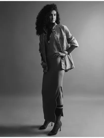 A powerful black and white full-length studio portrait. The strong composition and classic monochrome tones highlight the layered styling of a denim jacket over a long dress, demonstrating a timeless approach to fashion portraiture.