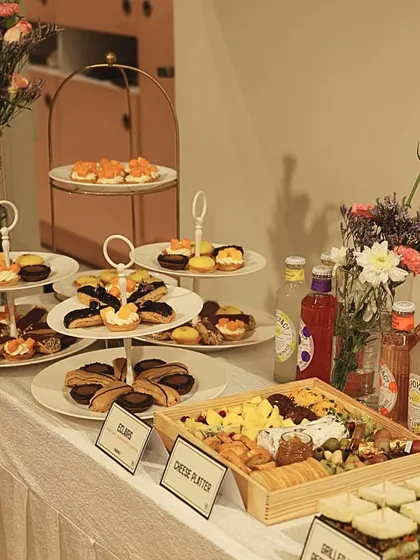 A full catering spread, featuring tiered stands of pastries, a cheese platter, mini sandwiches, and eclairs.