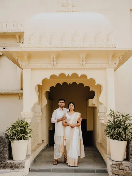 A serene and elegant portrait of the couple in beautiful white and gold traditional wear, standing in a classic archway.