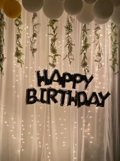 A beautiful room decoration featuring a wall of fairy lights behind a sheer white curtain. The decor is accented with hanging artificial vines and a black "Happy Birthday" foil balloon set.