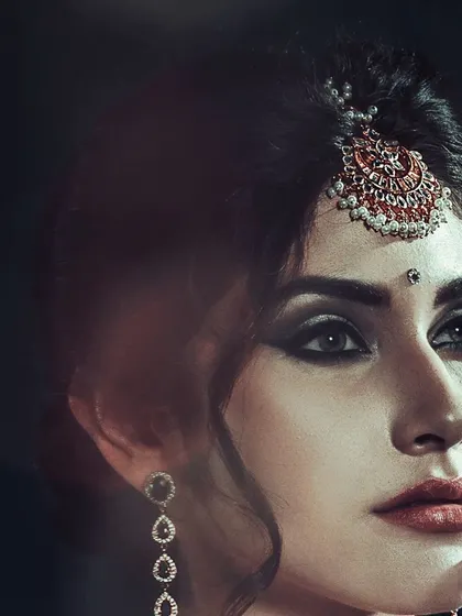A striking beauty shot with a focus on traditional Indian jewelry, including a maang tikka. The dark background and dramatic lighting create a moody and intense portrait.