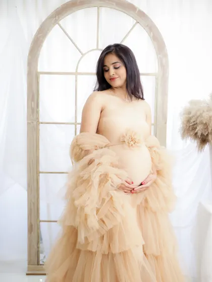 A delicate and ethereal portrait. The mom-to-be is dressed in a sheer, ruffled nude gown, standing by a window in a light-filled studio.