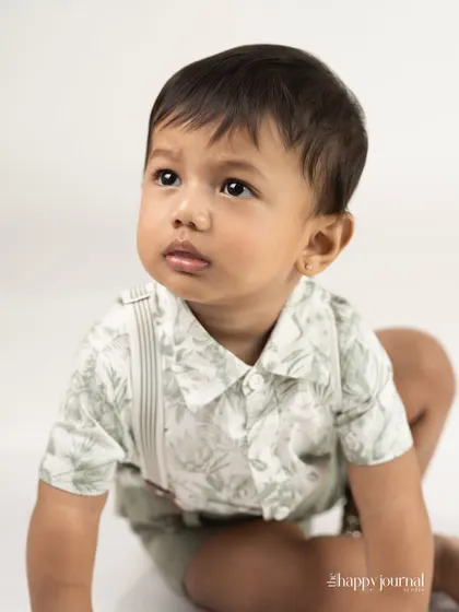 A baby boy in a stylish outfit with suspenders, looking up with wide, curious eyes. A classic and clean studio portrait.