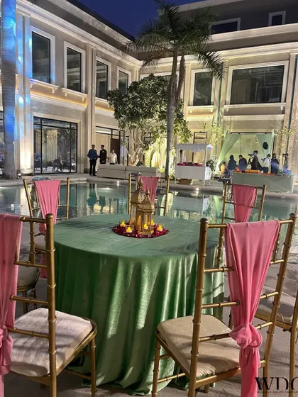 A charming poolside setup. The green tablecloth, pink chair drapes, and a golden lantern centerpiece with rose petals create an elegant and romantic atmosphere.