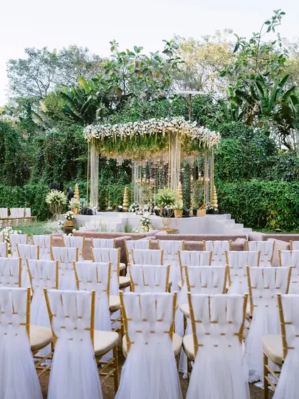 A serene outdoor mandap set against a backdrop of lush trees. The chairs, elegantly draped in white fabric, face the beautiful green and white floral structure.
