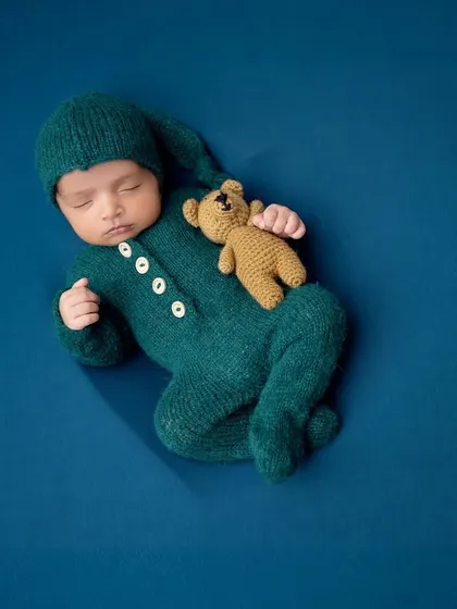 A sleeping baby in a teal sleeper and gnome hat, cuddling a tiny teddy bear. The solid blue background makes the colors stand out beautifully.