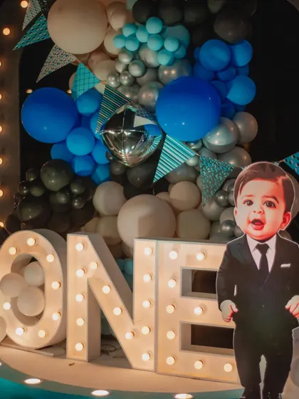 A "Boss Baby" themed first birthday, featuring large 'ONE' marquee lights and a life-sized cutout of the little boss himself. The blue, silver, and white balloon backdrop adds a celebratory touch.
