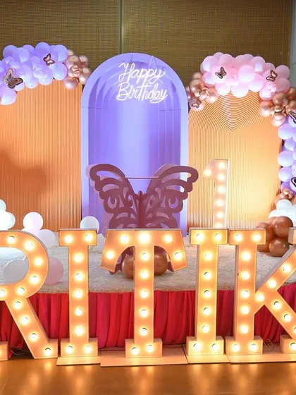 A first birthday party with a pastel butterfly theme. The setup includes large marquee letters, a purple lighted arch, and soft-colored balloon arrangements for a gentle and pretty look.
