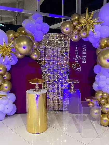 A luxurious birthday setup with a purple and gold theme. The decoration includes a shimmer panel, balloon arches, and golden pedestals.